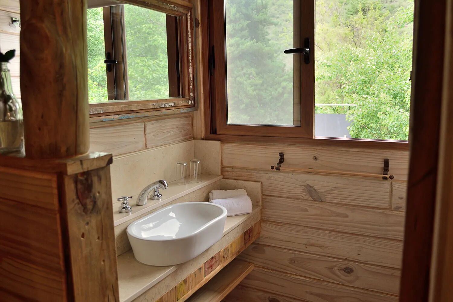 A wooden bathroom with a white sink, two faucets, and a mirror above it. There is a window with a view of greenery outside.