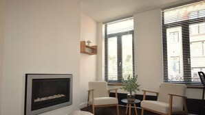 Un salon avec une cheminée, deux fauteuils, une petite table et une grande fenêtre avec des stores.