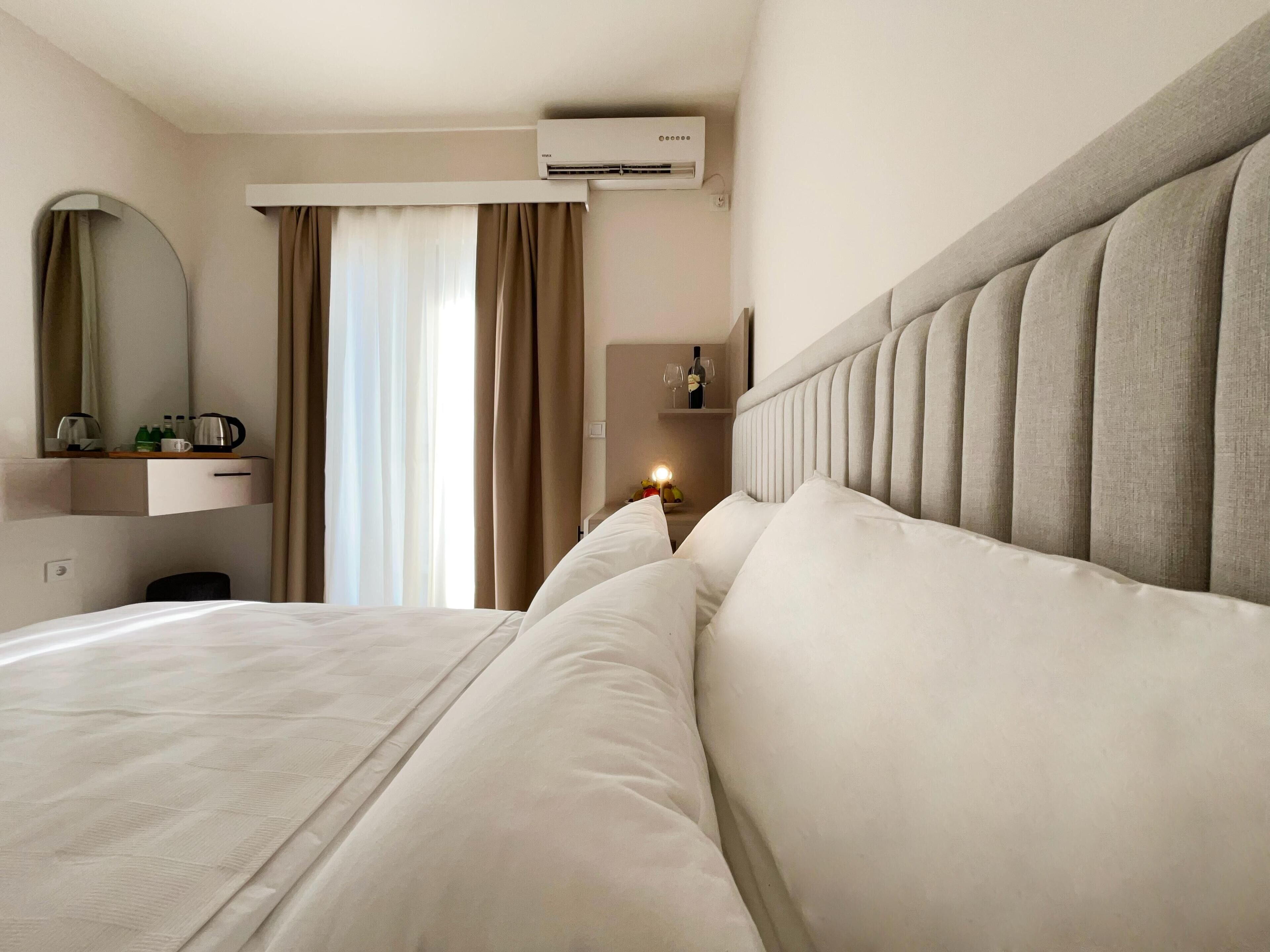 A hotel room with a large bed, a bedside table with a mirror, a minibar, and an air conditioning unit.