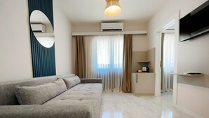 Una sala de estar moderna con un sofá gris, una pequeña cocina y un televisor de pantalla plana montado en la pared.