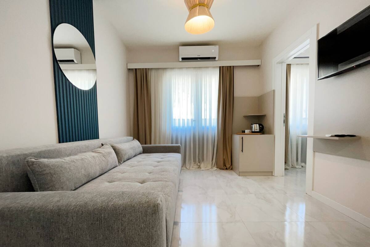 A modern living room with a grey sofa, a small kitchen area, and a flat-screen TV mounted on the wall.