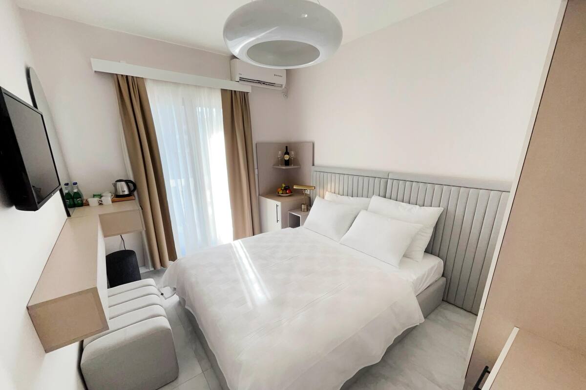 A modern hotel room with a large bed, a flat-screen TV mounted on the wall, a bedside table with a lamp, and a window with curtains.