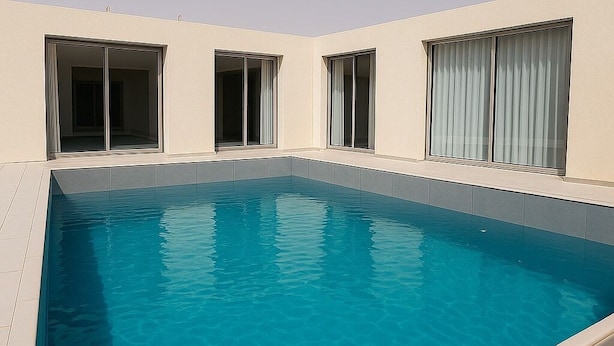 A rectangular swimming pool with clear blue water, surrounded by a light-colored deck and a beige building facade with large windows. - Royal Palace Compound (Riyadh)