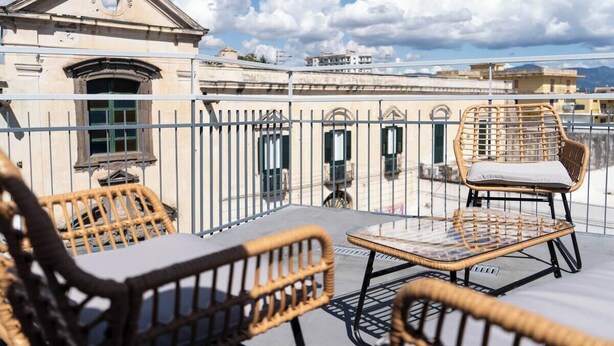 A rooftop terrace with wicker furniture, a small table, and a building with classical architecture in the background.