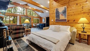 A cozy cabin-style room with a large bed, a sofa, a dining area, and a view of the forest through large windows. - The Robin's Nest, a cozy 3-bedroom cabin in Lake Harmony with Firepit, AC, WiFi (Lake Harmony)