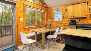 A wooden cabin interior with a dining table, white chairs, a kitchen area, and large windows overlooking a forest. - The Robin's Nest, a cozy 3-bedroom cabin in Lake Harmony with Firepit, AC, WiFi (Lake Harmony)