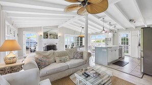 A spacious living room with a sofa, a coffee table, and a fireplace. There is a kitchen area with a refrigerator and a dining area visible through the glass doors.