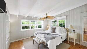 A bedroom with a large bed, a ceiling fan, a window with a view of greenery, a bedside table with a lamp, and a small bench.