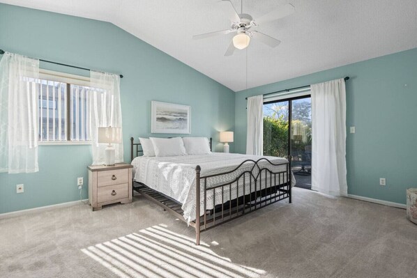 Une chambre à coucher avec un lit, une table de chevet, un ventilateur de plafond et une vue sur l’extérieur, visible à travers une porte vitrée.