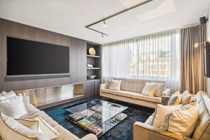 Een moderne woonkamer met een grote flatscreen televisie, een zithoek, een salontafel met boeken en een groot raam met gordijnen.