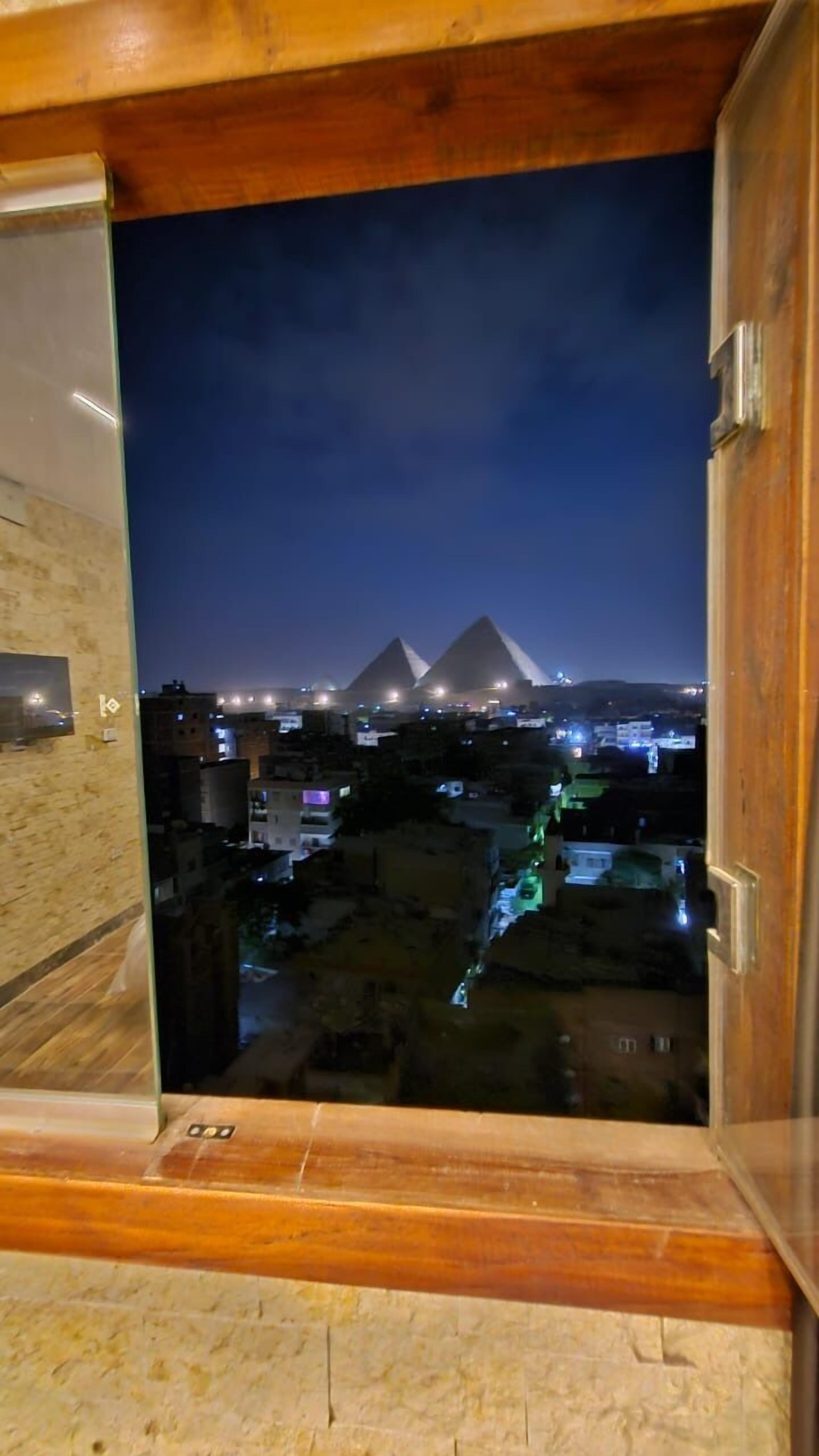 A view from a window overlooking a cityscape at night, featuring illuminated pyramids and surrounding buildings.