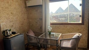 A room with a view of pyramids, a small table with chairs, a refrigerator, and a window with curtains. - Vida Pyramids Inn (Giza)