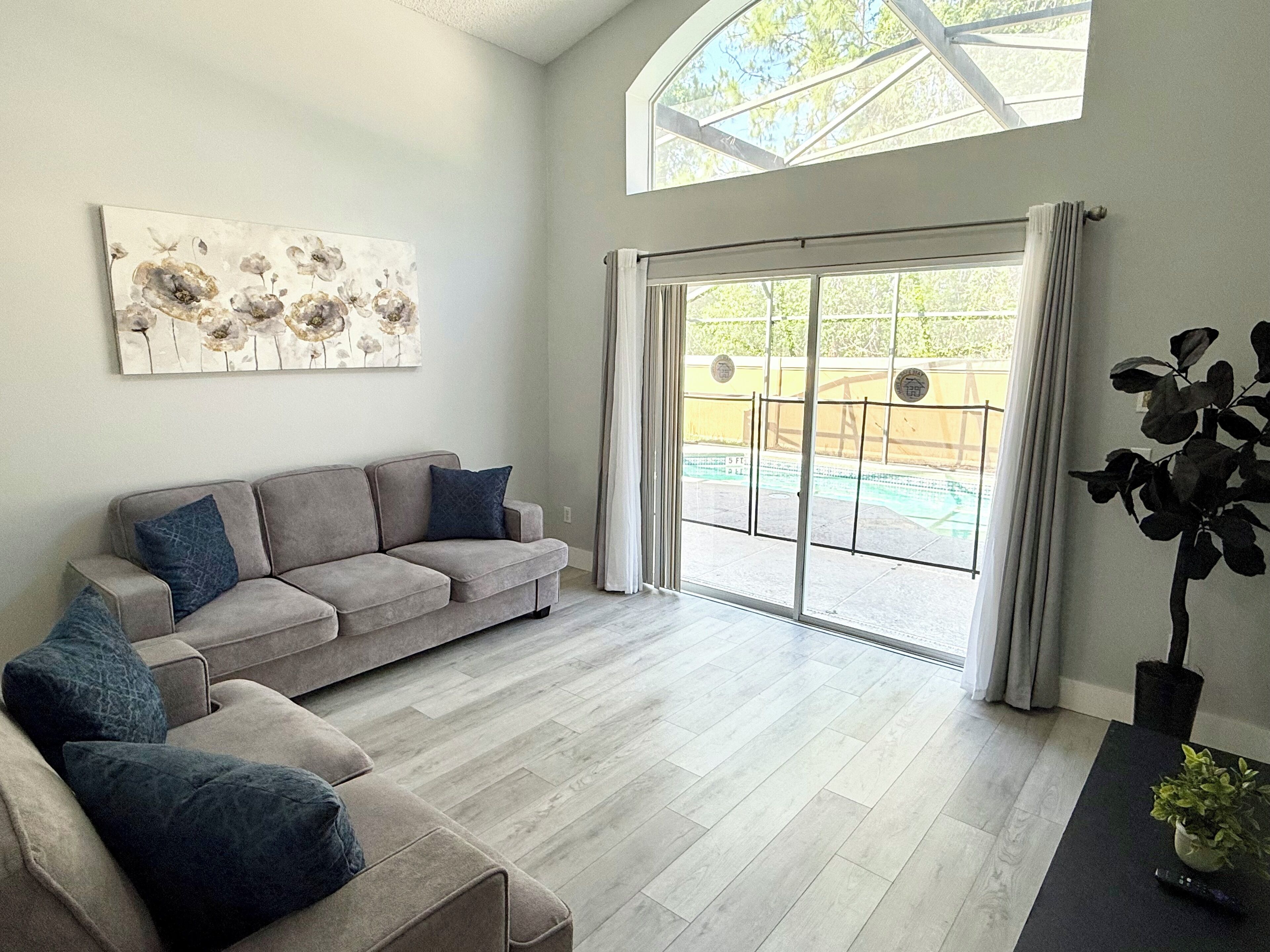 A spacious living room with a grey sofa, a coffee table, and a large window with a view of a swimming pool.