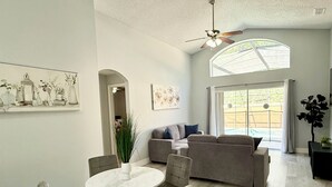 A spacious living room with a grey sofa, a round dining table with chairs, a ceiling fan, and a large window with a view of a pool. - Luxury Florida Villa with Private Pool - Free Heating (Kissimmee)