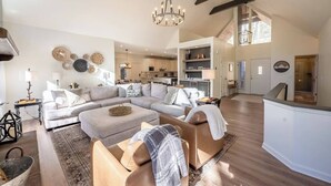 A spacious living room with a large sectional sofa, a coffee table, and a chandelier. There is a kitchen in the background with modern appliances and a dining area.