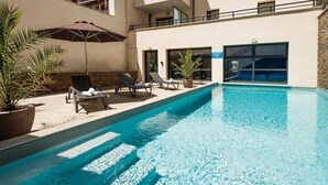 Ein Schwimmbecken mit Liegestühlen und Sonnenschirmen, umgeben von Kübelpflanzen und einem Gebäude mit großen Fenstern.