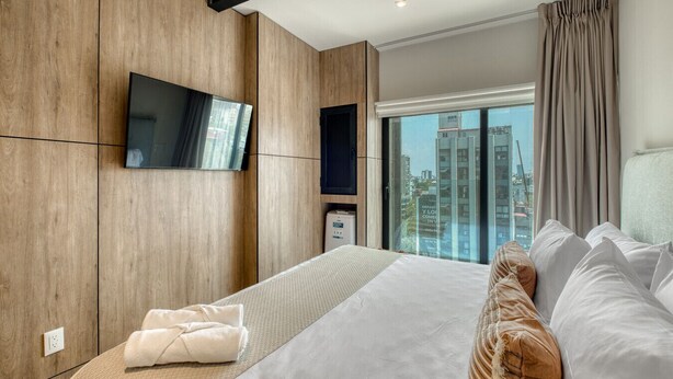 A modern hotel room with a large bed, a flat-screen TV mounted on the wall, a view of the city through the window, and a small bedside table.
