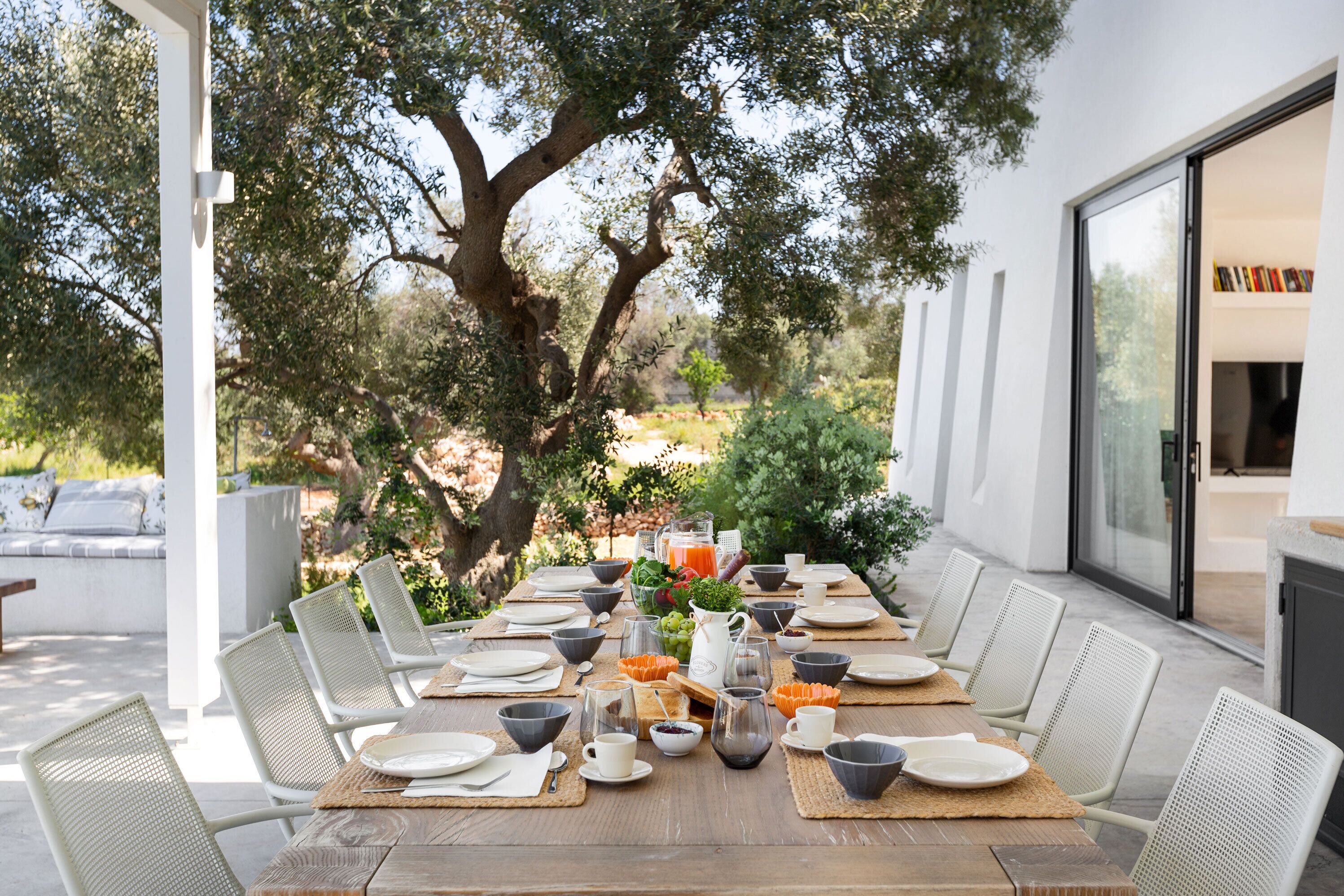 An outdoor dining area with a long wooden table set for a meal, surrounded by white chairs, with a large tree and a modern building in the background.