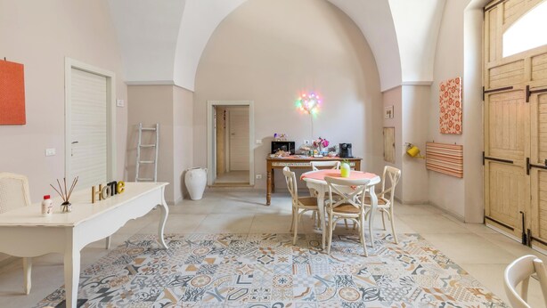 A dining area with a white table, chairs, and a patterned rug. There is a kitchen area in the background with a microwave and a coffee machine. - D.Home B&B - Camera quadrupla (Oria)