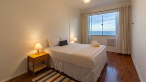 A bedroom with a bed, two bedside lamps, a ceiling fan, a window with curtains, and a view of the sea.