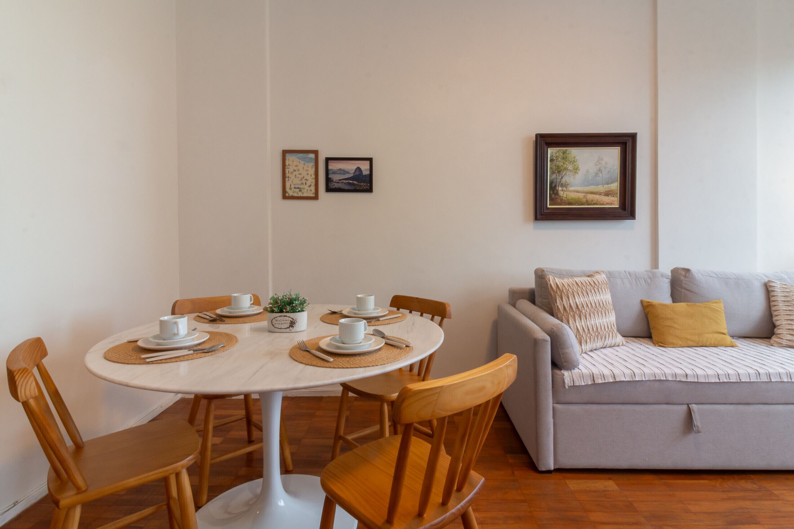 Un comedor con una mesa redonda, cuatro sillas de madera y un sofá gris con un cojín amarillo. Hay dos cuadros en la pared.