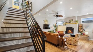 Una escalera con terminación en madera y barandal negro, que conduce a una sala de estar con chimenea, sofá y televisor montado en la pared.