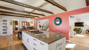 Una cocina amplia con una isla grande, vigas de madera en el techo y una distribución luminosa y abierta que da a una sala de estar con televisor y asientos.