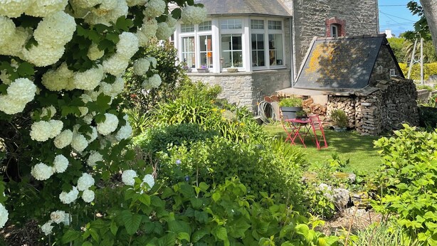 A stone house with a large garden, a tree with white flowers, and a clear blue sky.