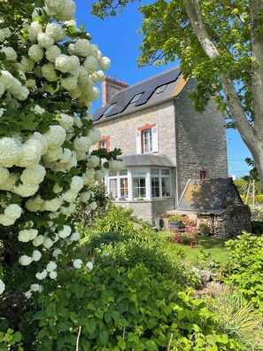 A stone house with a large garden, a tree with white flowers, and a clear blue sky. - Charming house 400 m from the beach (Saint-Cast-le-Guildo)