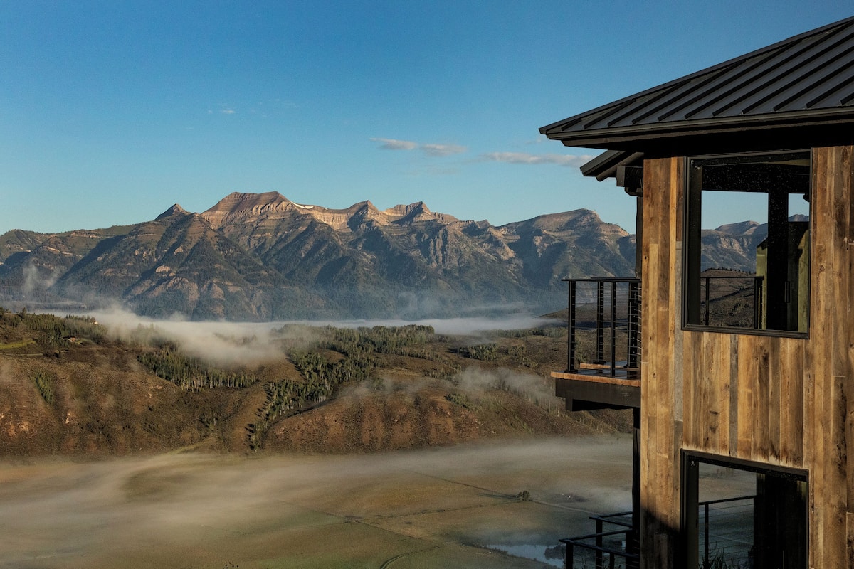 A wooden cabin with a balcony overlooking a mountainous landscape with visible mist.