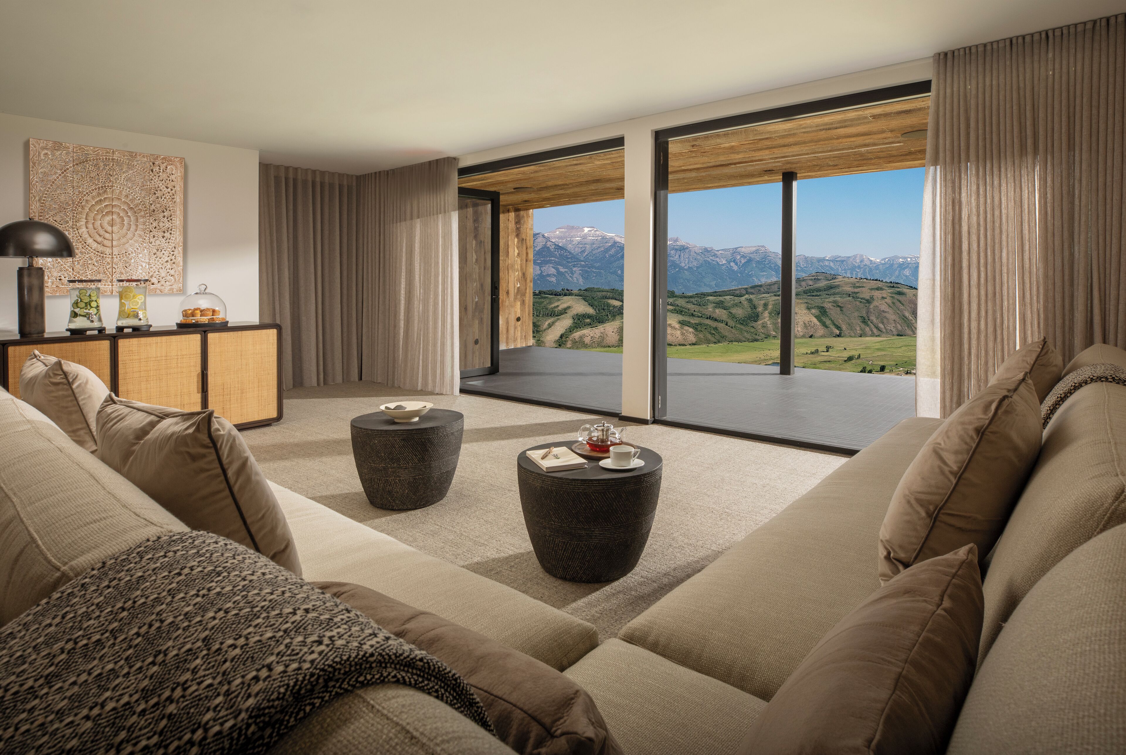 A modern living room with a large sofa, two round side tables, and a view of mountains through floor-to-ceiling windows.