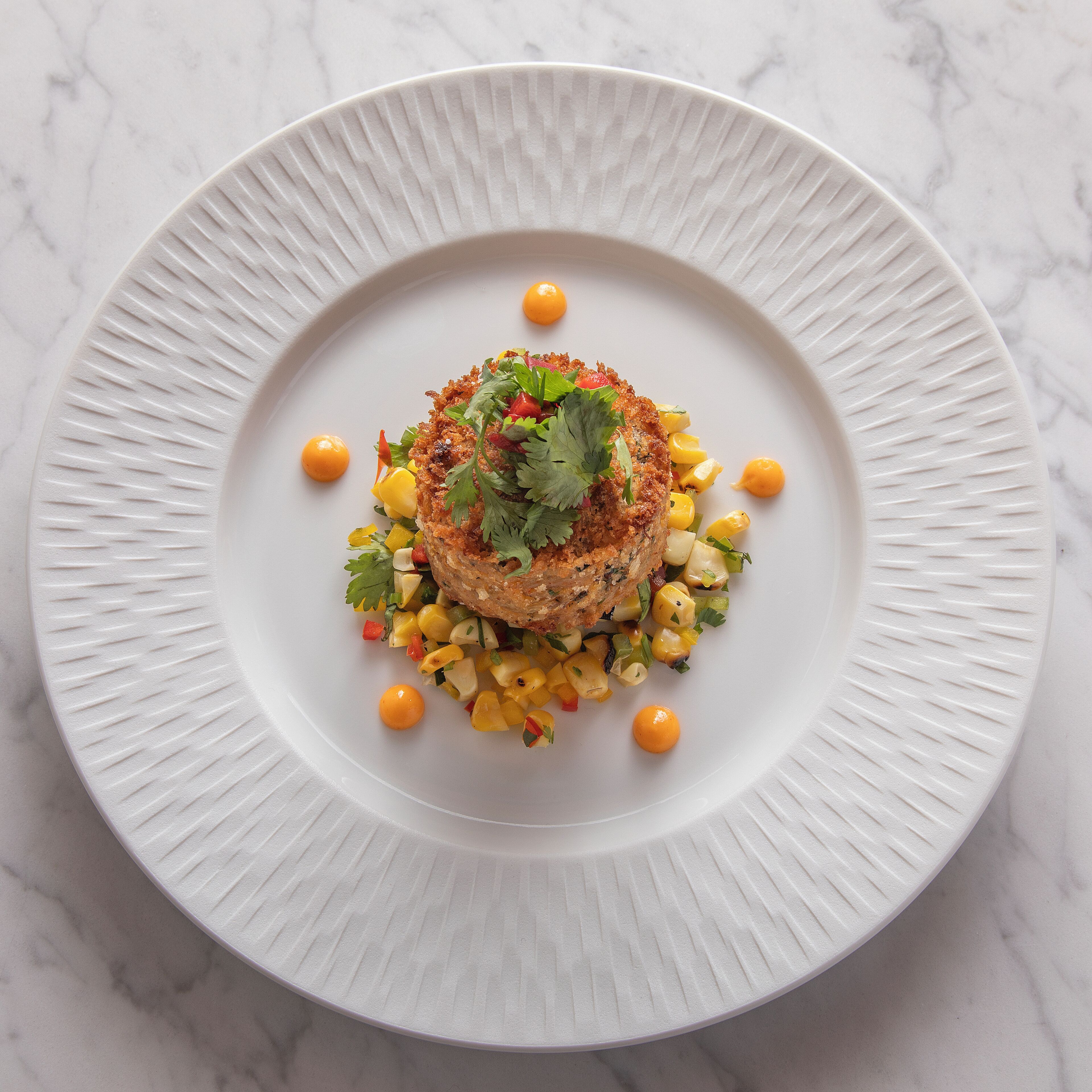 A white plate with a textured edge, containing a central food item surrounded by diced vegetables and small yellow spheres.