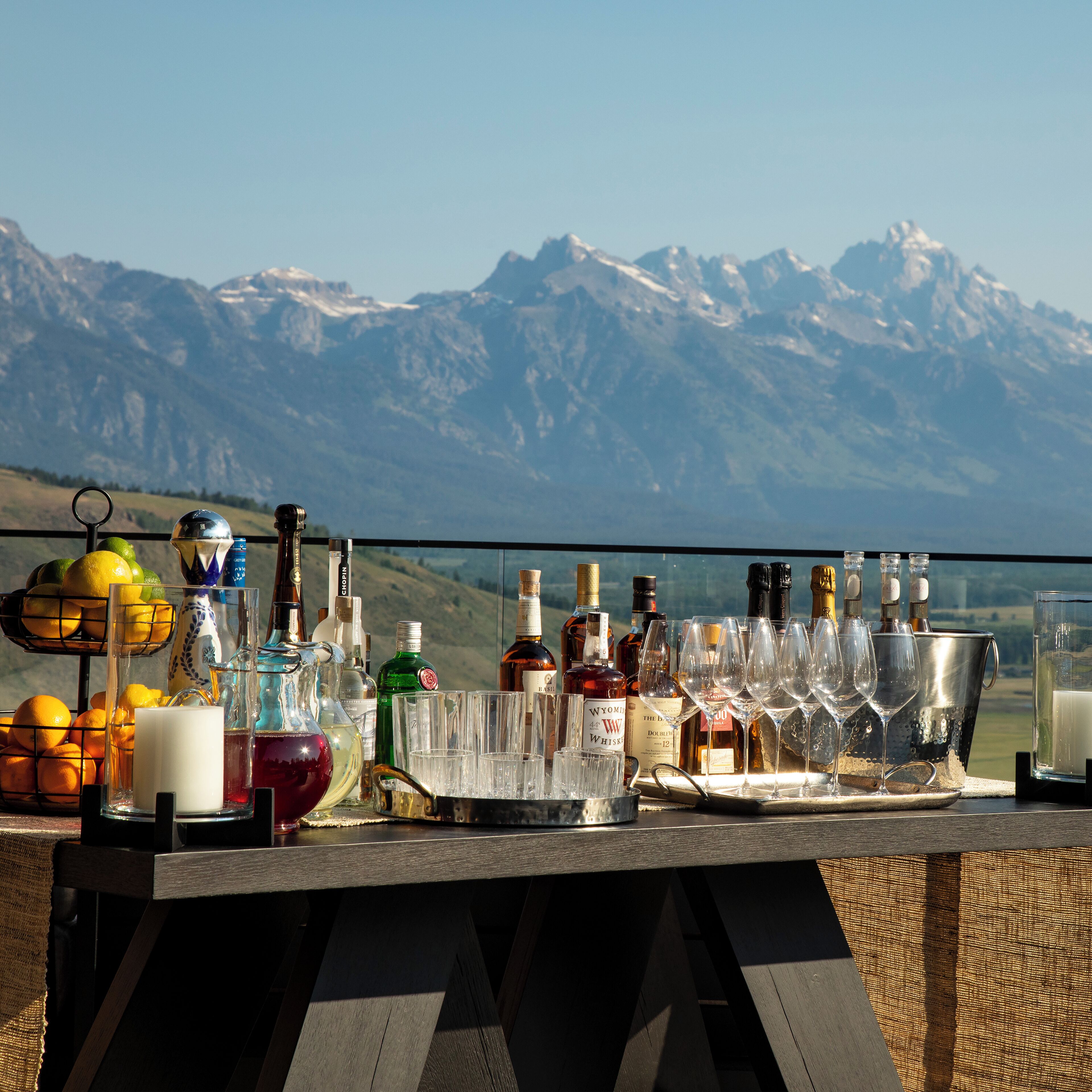 Un bar avec différentes bouteilles et différents verres, un saladier rempli d’oranges et une bougie posés sur une table, avec une vue sur les montagnes en arrière-plan.