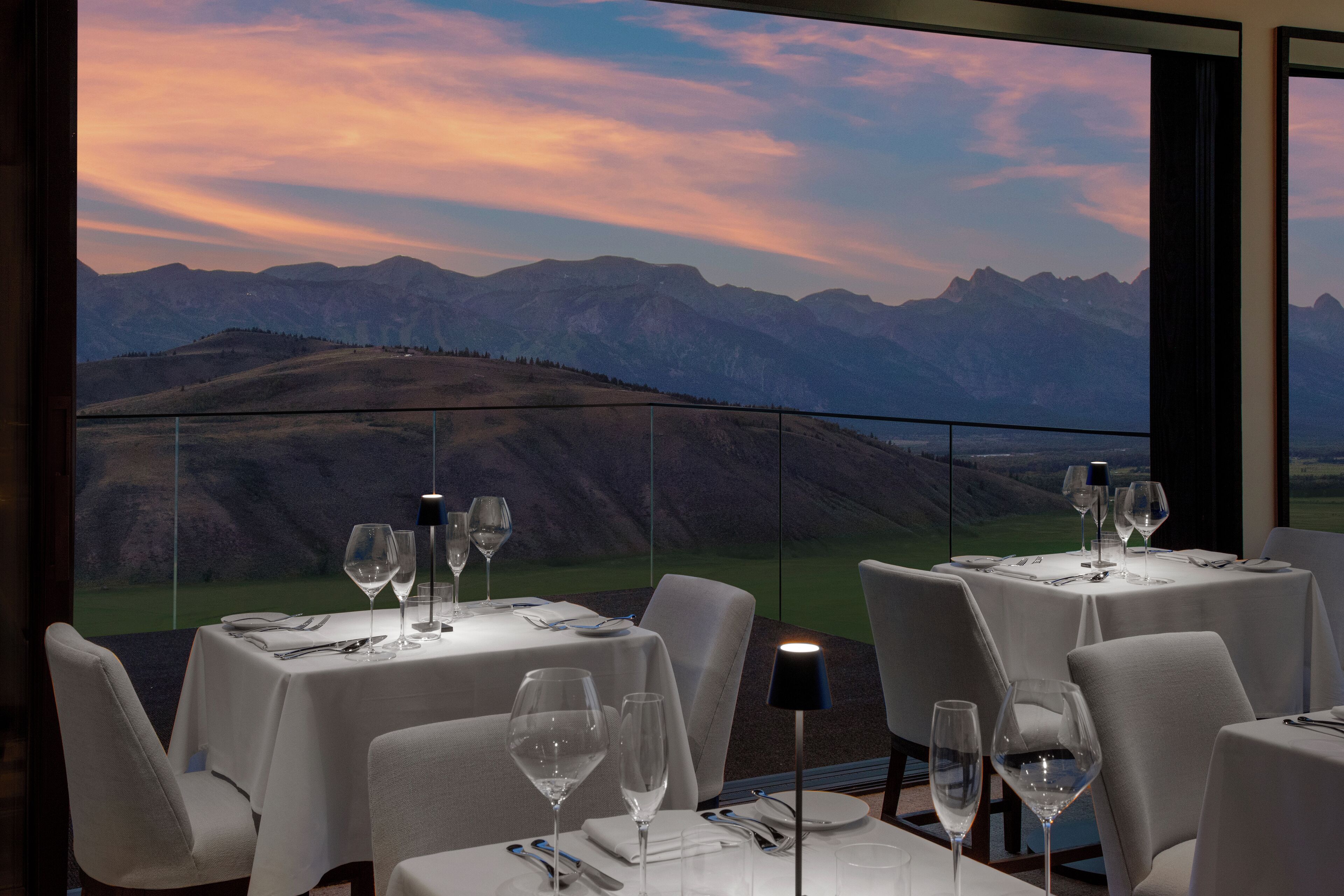 Uma área de refeições com mesas preparadas para dois, cada uma com uma taça de vinho, talheres e uma luminária. O espaço possui grandes janelas com vista para montanhas.
