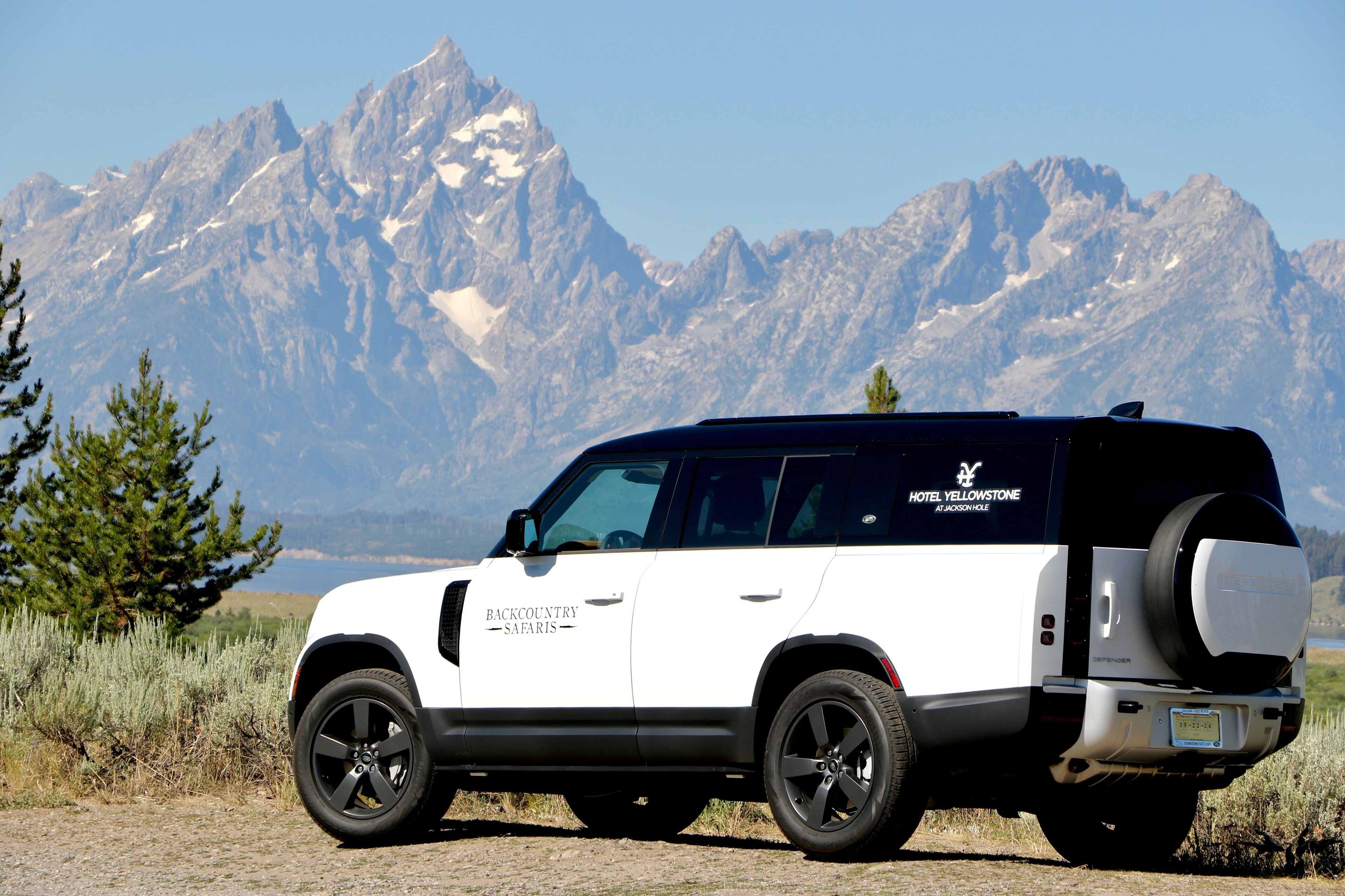 A white SUV with "Backcountry Safaris" and "Hotel Yellowstone" branding parked in a natural setting with mountains in the background.