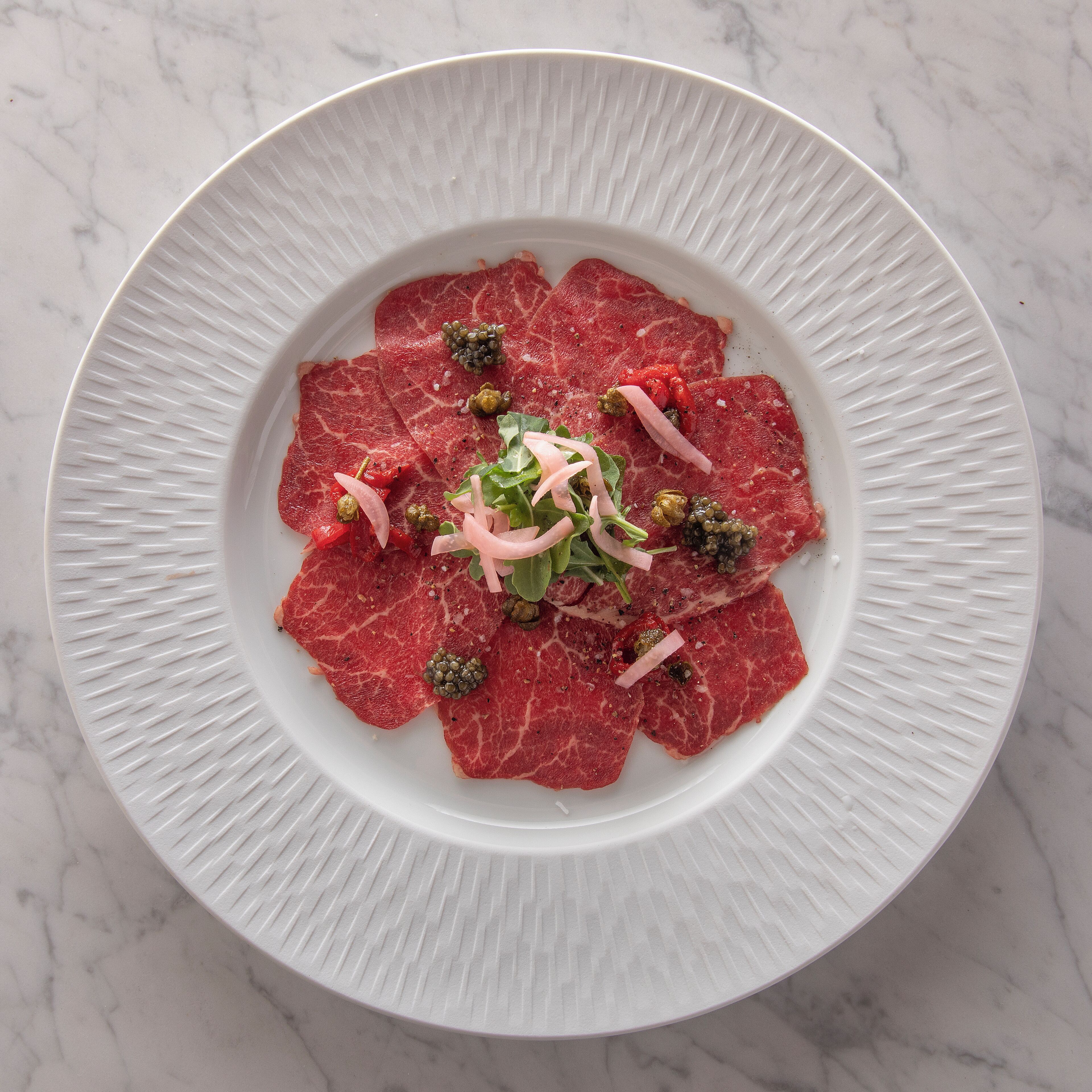 A white plate with thinly sliced red meat, garnished with green capers, red onion, and green leaves.