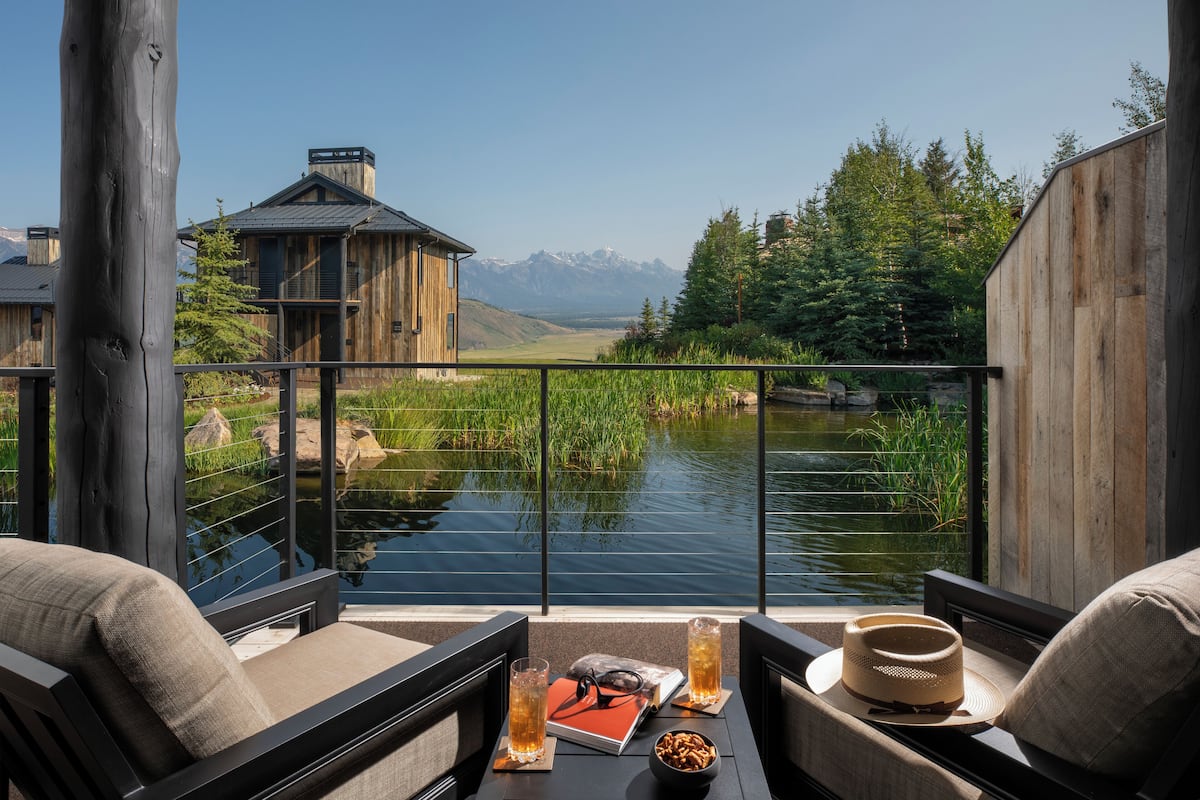A view from a balcony overlooking a river, with a wooden building in the background and a mountain range in the distance.