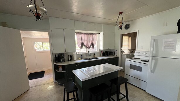 Cocina compacta con refrigerador, estufa y microondas blancos. Cuenta con una pequeña mesa de comedor y dos taburetes. Se ve una ventana con cortinas, y una puerta conduce a otra habitación.
