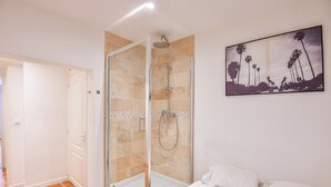 A hotel room with a glass-enclosed shower, a bed with white linens, and a painting of palm trees on the wall.