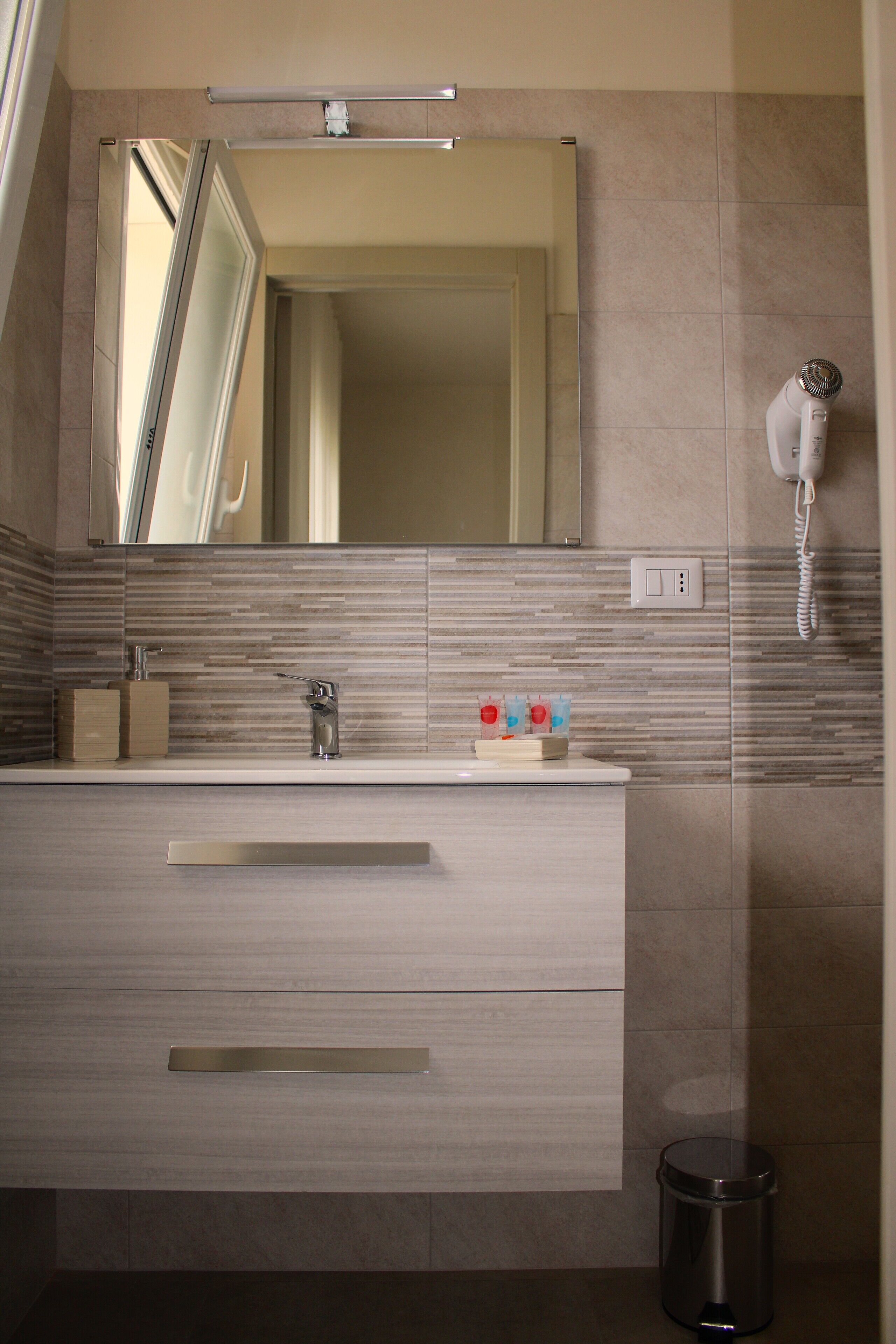 A modern bathroom with a large mirror, a sink with a faucet, a trash bin, and a hairdryer on the wall.