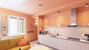 A modern kitchen with a built-in oven, microwave, and sink. There is a yellow sofa with a red cushion in the adjacent living area. - Materia Terra&Vidru Studio by Propertize (Cagliari)