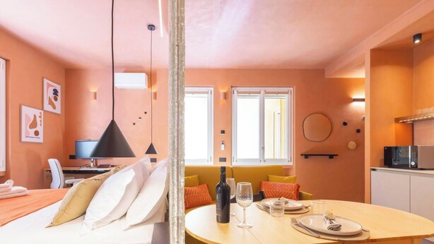 A modern hotel room with a bed, a dining table set for two, a kitchen area, and a view of the outside. - Materia Terra&Vidru Studio by Propertize (Cagliari)
