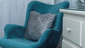 Un sillón azul aguamarina con patas de madera y un cojín estampado, ubicado junto a un aparador blanco con cajones.