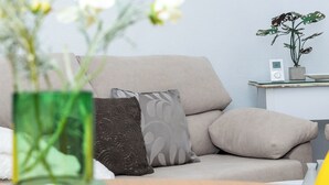 Una sala de estar con un sofá color beige, un jarrón verde con flores, un cuadro en la pared y una mesita con una planta y un altavoz.
