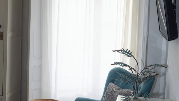 Un dormitorio con una cama, un sillón azul, una mesita redonda pequeña, un armario blanco y una planta en maceta.