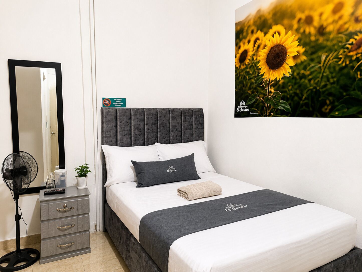 Un dormitorio con cama, mesita de noche, espejo, ventilador y un cuadro de girasoles colgado en la pared.