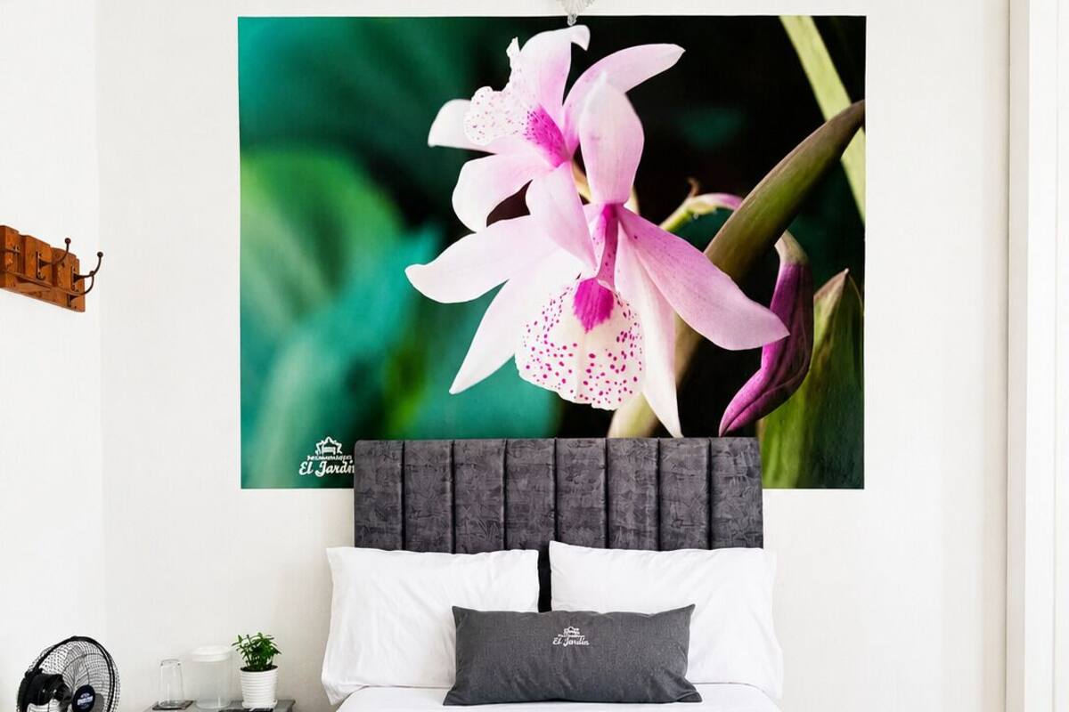 Une chambre à coucher comprenant un lit, une table de chevet, un ventilateur, un oreiller, une couverture et un tableau représentant une orchidée rose, accroché au mur.