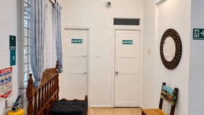A room with a bed, two doors marked 'BAÑO,' a fire extinguisher, a mirror, and a chair with a picture on the backrest. - Hospedajes El Jardin (Envigado)