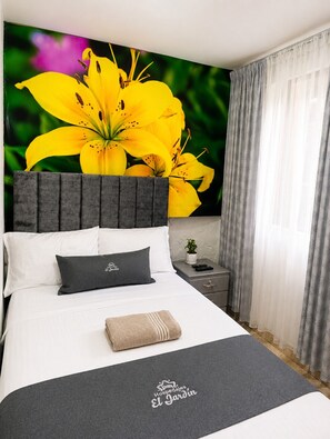 Habitación de hotel con una cama grande, cabecera gris, manta gris, almohada gris, una mesita de noche con una planta y una ventana con cortinas grises.