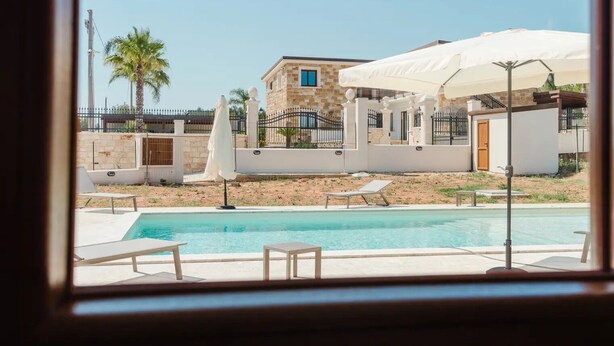 A pool area with lounge chairs and umbrellas, a stone building, and a fenced property. - VILLA CON PISCINA (Balestrate)